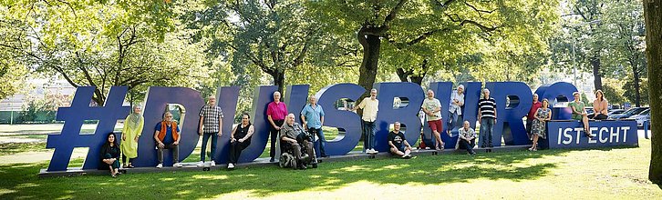 Das Team der Selbsthilfe-Kontaktstelle Duisburg mit Kolleg*innen unter Bäumen im großen Duisburg-Schriftzug "#Duisburg ist echt"